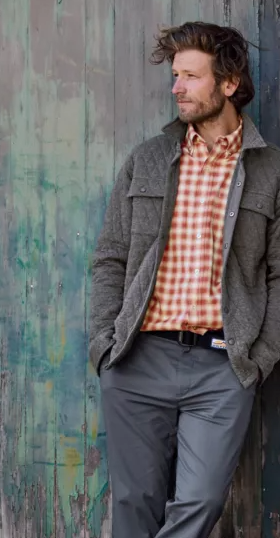 A man leaning against a wall wearing an Orvis plaid shirt and overcoat with workwear pants
