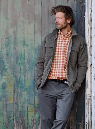 A man leaning against a wall wearing an Orvis plaid shirt and overcoat with workwear pants