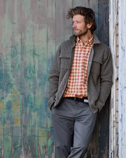 A man leaning against a wall wearing an Orvis plaid shirt and overcoat with workwear pants