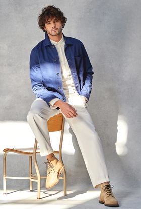 A man sitting on a chair while wearing a Todd Snyder outfit with a denim overcoat and cream pants
