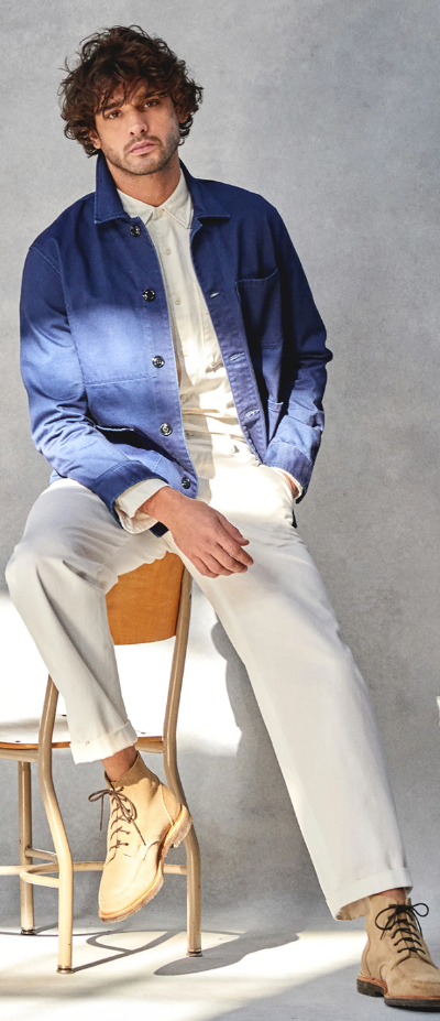A man sitting on a chair while wearing a Todd Snyder outfit with a denim overcoat and cream pants