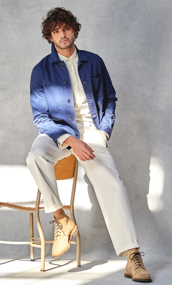 A man sitting on a chair while wearing a Todd Snyder outfit with a denim overcoat and cream pants