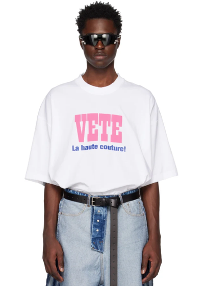 A man wearing a Vetements outfit featuring a white VETE t-shirt and blue jeans