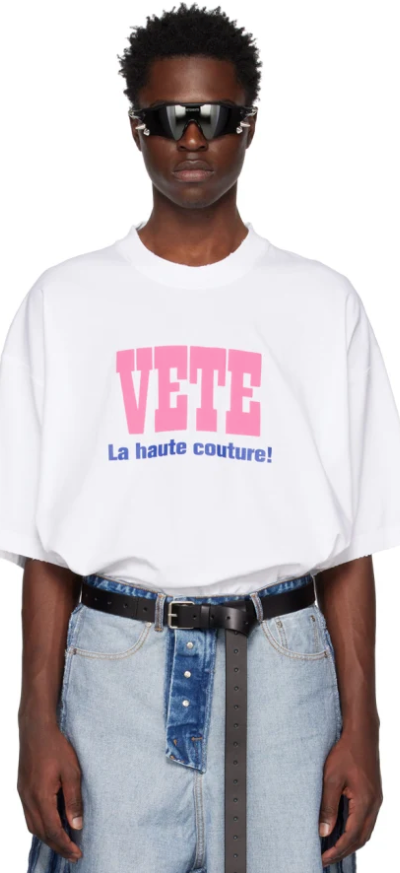 A man wearing a Vetements outfit featuring a white VETE t-shirt and blue jeans