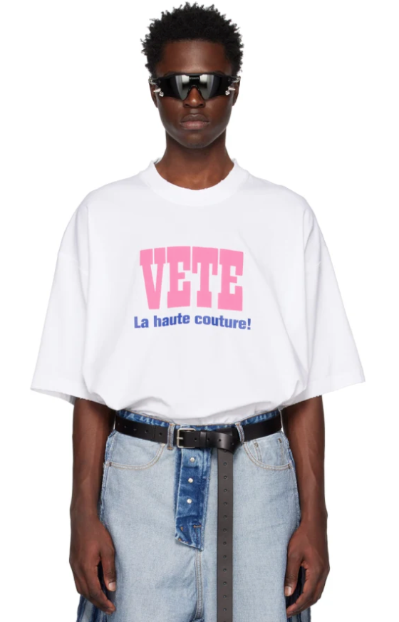 A man wearing a Vetements outfit featuring a white VETE t-shirt and blue jeans