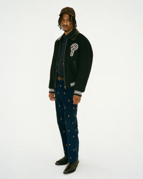 A man wearing a Patta coaches jacket with navy blue pants and black pointed dress shoes
