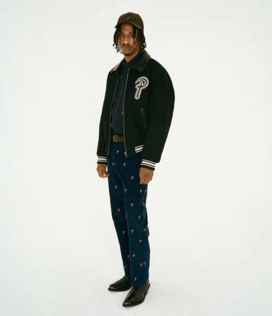 A man wearing a Patta coaches jacket with navy blue pants and black pointed dress shoes