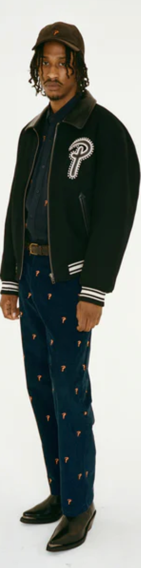 A man wearing a Patta coaches jacket with navy blue pants and black pointed dress shoes
