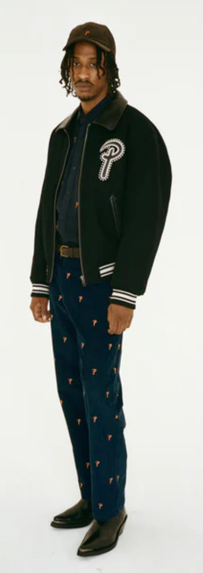 A man wearing a Patta coaches jacket with navy blue pants and black pointed dress shoes