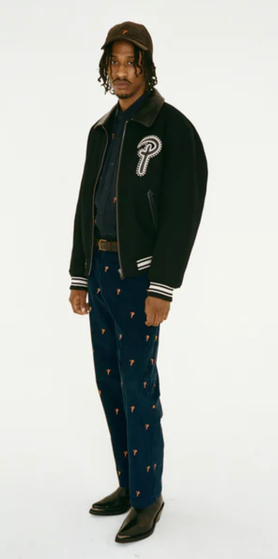 A man wearing a Patta coaches jacket with navy blue pants and black pointed dress shoes