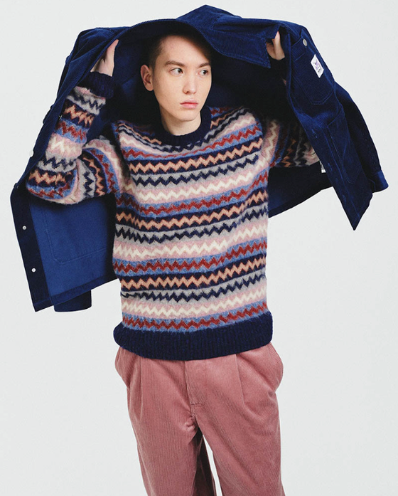 A man wearing a striped Howlin wool sweater, a navy blue overcoat and red pants