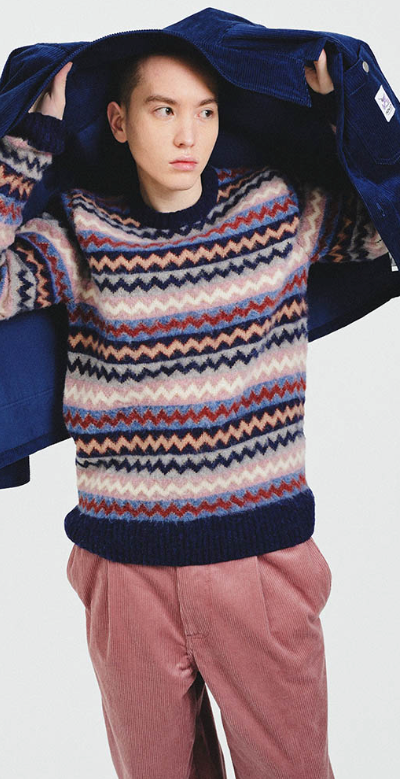 A man wearing a striped Howlin wool sweater, a navy blue overcoat and red pants