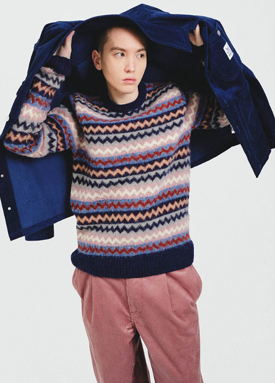 A man wearing a striped Howlin wool sweater, a navy blue overcoat and red pants