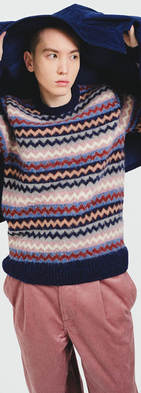 A man wearing a striped Howlin wool sweater, a navy blue overcoat and red pants