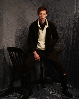 A man wearing a dark Mr. Oliver jacket, beige zip sweater with black pants and boots