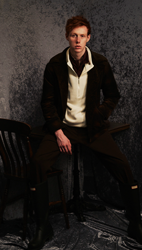 A man wearing a dark Mr. Oliver jacket, beige zip sweater with black pants and boots