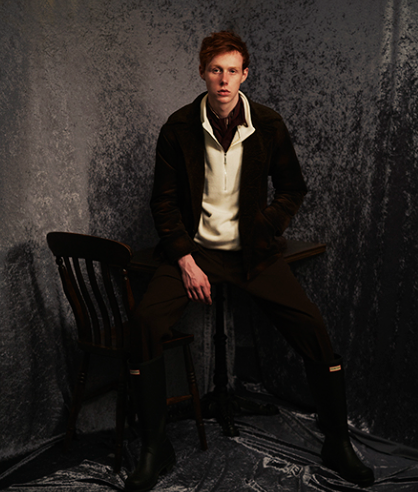 A man wearing a dark Mr. Oliver jacket, beige zip sweater with black pants and boots