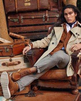 A man sitting in a chair wearing a Banana Republic trench coat, a brown jacket underneath and brown shoes