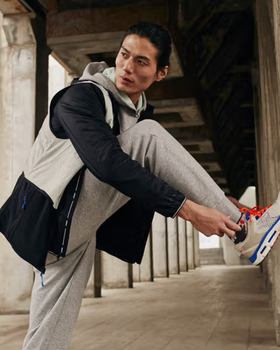 A man tying up his Under Armor shoes while he wears an Under Armor grey sweatpants and a black and grey jacket