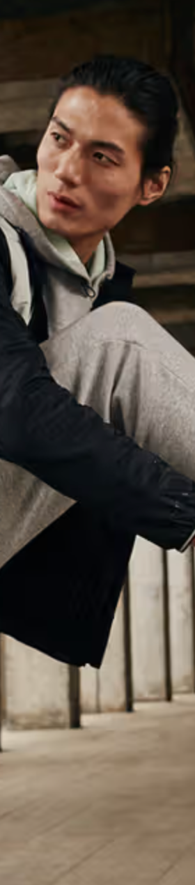 A man tying up his Under Armor shoes while he wears an Under Armor grey sweatpants and a black and grey jacket