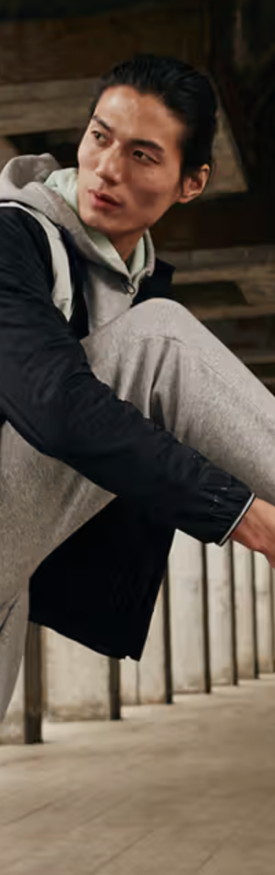 A man tying up his Under Armor shoes while he wears an Under Armor grey sweatpants and a black and grey jacket