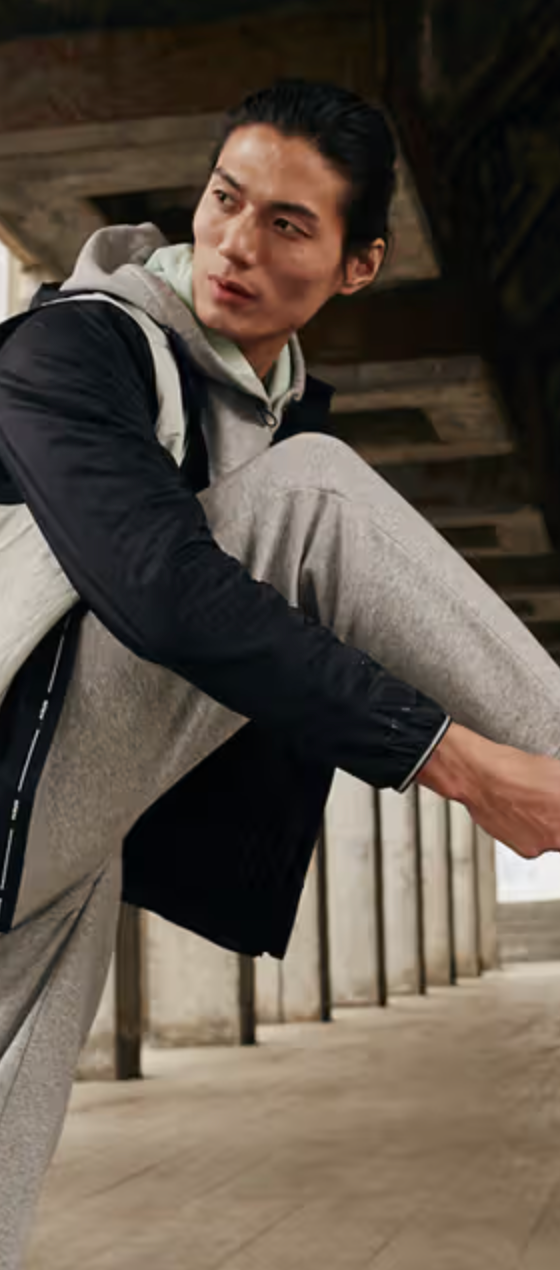 A man tying up his Under Armor shoes while he wears an Under Armor grey sweatpants and a black and grey jacket