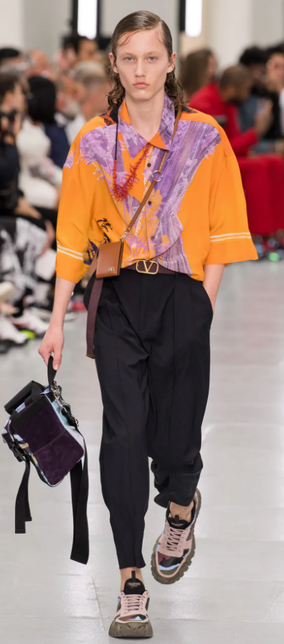 A person wearing an orange Valentino button up, black pants and colorful sneakers
