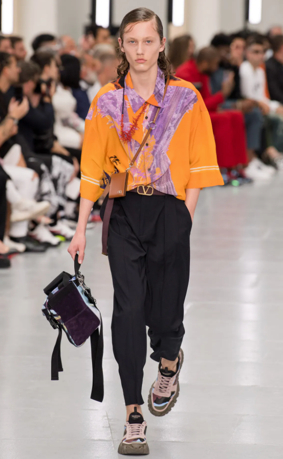 A person wearing an orange Valentino button up, black pants and colorful sneakers