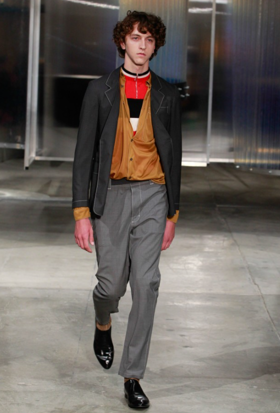 A man wearing a Prada outfit with grey pants and a brown cardigan