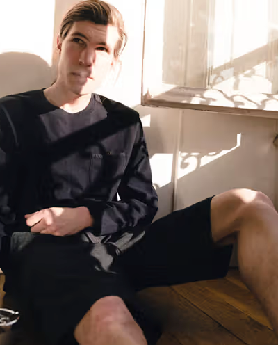 A man sitting in the sun wearing a Magenta Skateboards long-sleeve and shorts