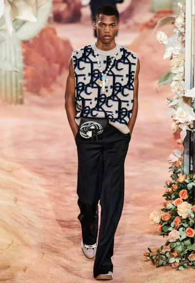 A man wearing a Dior allover print vest, black pants and a colorful cross necklace