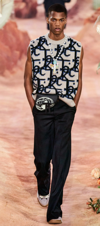 A man wearing a Dior allover print vest, black pants and a colorful cross necklace