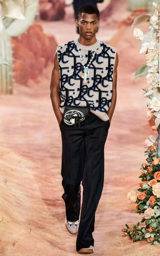 A man wearing a Dior allover print vest, black pants and a colorful cross necklace