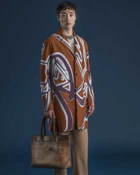 A man wearing a long Berluti jacket with a large purple pattern on top and brown pants