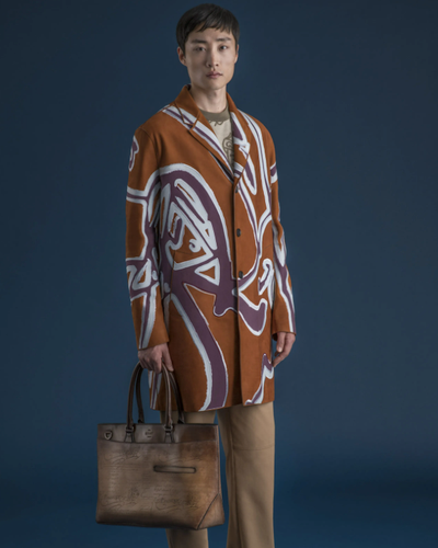 A man wearing a long Berluti jacket with a large purple pattern on top and brown pants