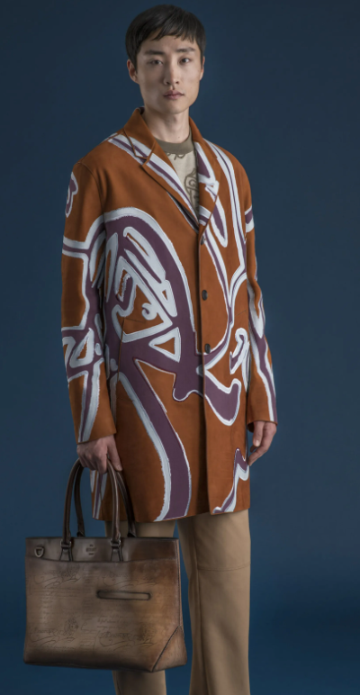 A man wearing a long Berluti jacket with a large purple pattern on top and brown pants