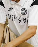 A close up of a man wearing a white Welcome Skateboards and Adidas collaboration soccer style jersey