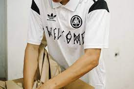 A close up of a man wearing a white Welcome Skateboards and Adidas collaboration soccer style jersey