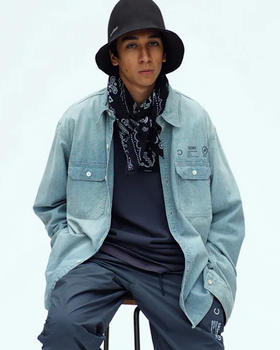 A man sitting in a Neighborhood outfit feating a navy bandana, denim overshirt, technical pants and a bucket hat