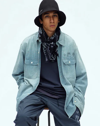 A man sitting in a Neighborhood outfit feating a navy bandana, denim overshirt, technical pants and a bucket hat