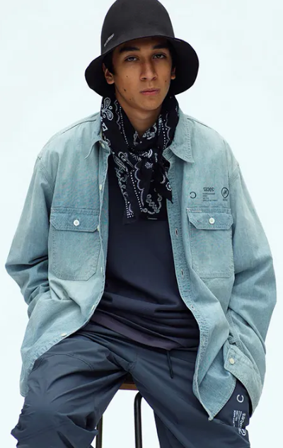 A man sitting in a Neighborhood outfit feating a navy bandana, denim overshirt, technical pants and a bucket hat