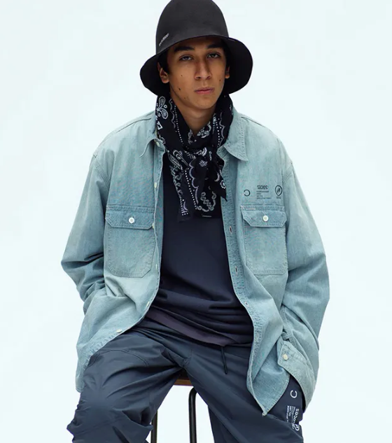A man sitting in a Neighborhood outfit feating a navy bandana, denim overshirt, technical pants and a bucket hat