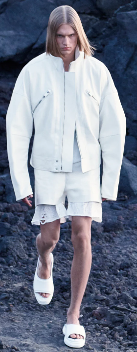 A man walking outdoors wearing an all-white Alyx jacket, shorts and slippers