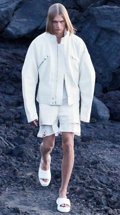 A man walking outdoors wearing an all-white Alyx jacket, shorts and slippers