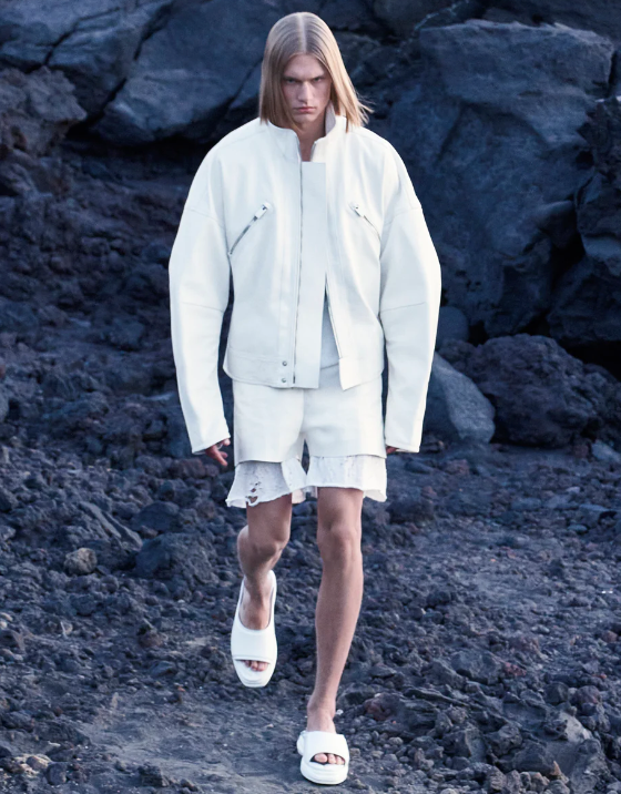 A man walking outdoors wearing an all-white Alyx jacket, shorts and slippers