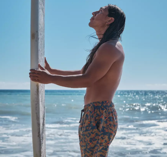 A man standing with a surf-board with colorful print swim shorts by PrAna