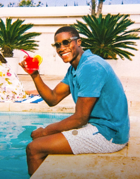 A man sitting by a pool in Ted Baker swimshorts and a blue polo
