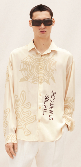 A man wearing a Jacquemus outfit featuring a cream button-up, white pants and modern sunglasses