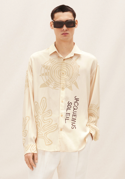 A man wearing a Jacquemus outfit featuring a cream button-up, white pants and modern sunglasses