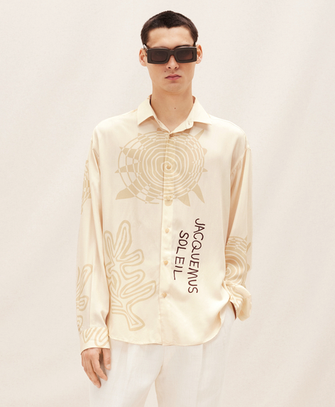 A man wearing a Jacquemus outfit featuring a cream button-up, white pants and modern sunglasses